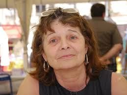 Photo couleur portrait de Claire Auzias, morte le 6 août 2024. Elle porte des boucles d'oreilles rondes et des lunettes sur la tête. Elle a un t-shirt sans manche noir et un petit sourire en coin.