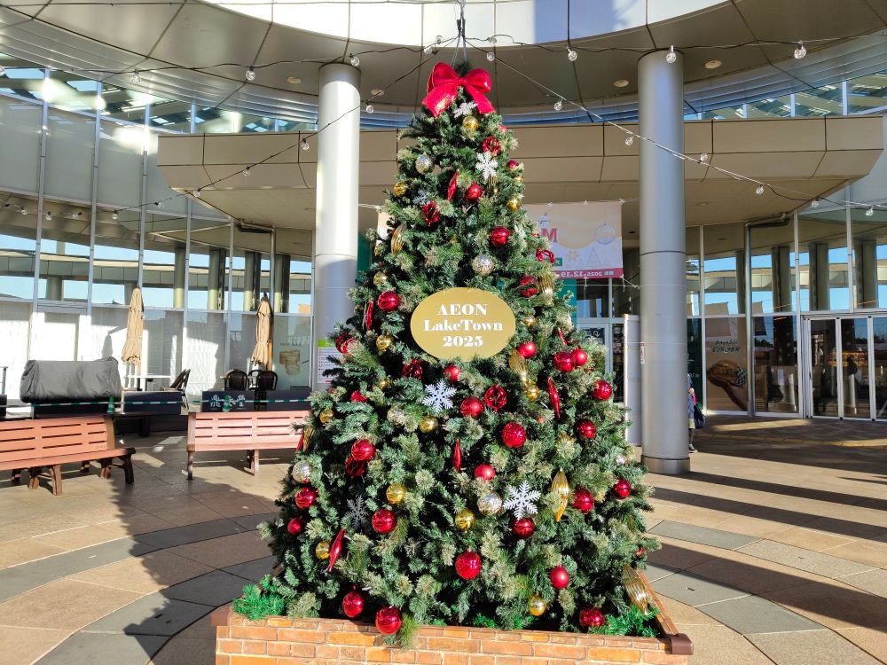 昼間に撮ったクリスマスツリー。赤くて丸いオーナメントがメイン。てっぺんに赤いリボンが付けてある。真ん中にあるゴールドの楕円のプレートにイオンレイクタウン2025と書かれている。