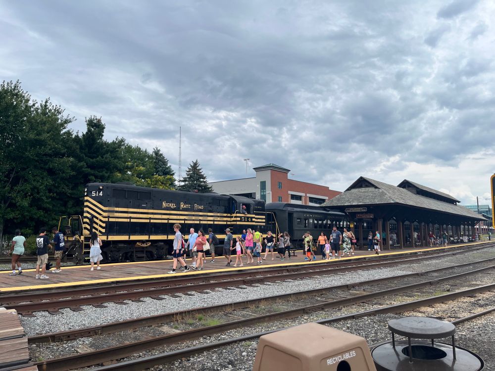 A black steam engine with passenger cars sits at the station. A group of people walk along side it as they disembark 