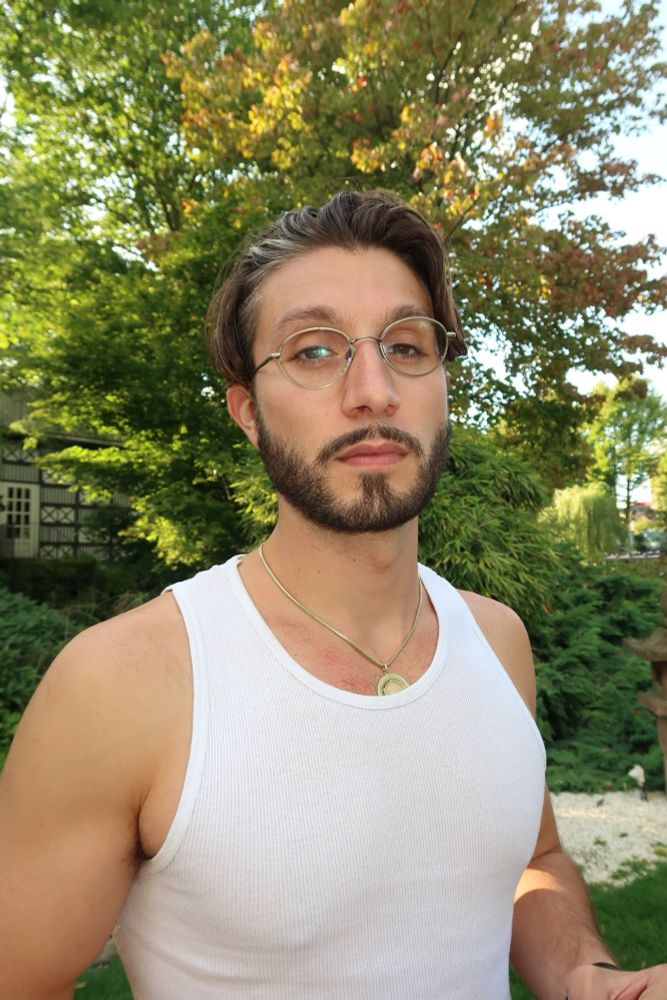 Bbno$, a man wearing a white tank top, gold necklace, and gold circular glasses; he has brown hair with a white streak toward the front and a beard