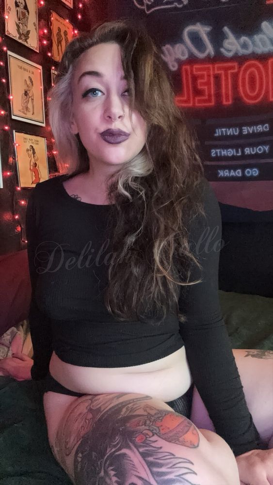 Delilah sitting on a green bedspread wearing a black long sleeve shirt and black underwear; her brown hair has a blonde streak and she is wearing purple lipstick 