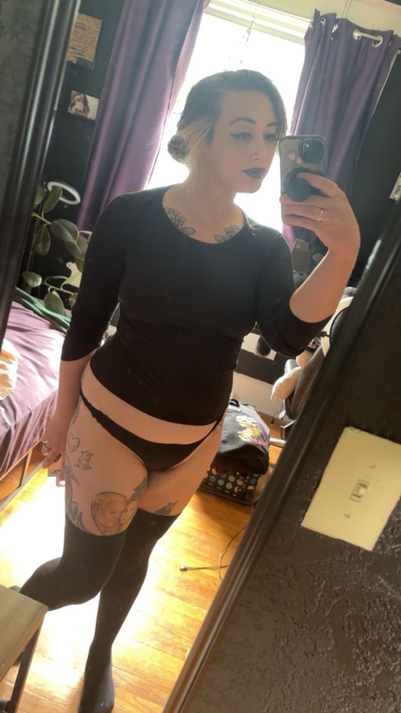 Delilah in a mirror selfie wearing black underwear, a black long sleeves shirt, and black thigh high socks; her hair is up in a bun and she’s wearing black eyeliner and purple lipstick