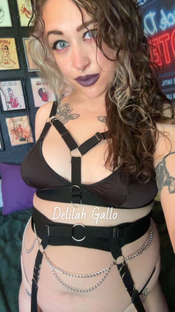 Delilah in a black bra with a harness. A waist chain belt with garters sitting on a bed 