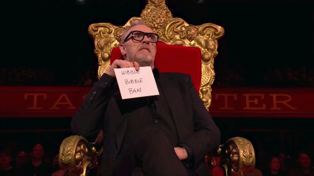 Greg Davies on his Taskmaster throne, with a very serious expression on his face as he holds up a card that says, "WIBBLE. BIBBLE. BAM."