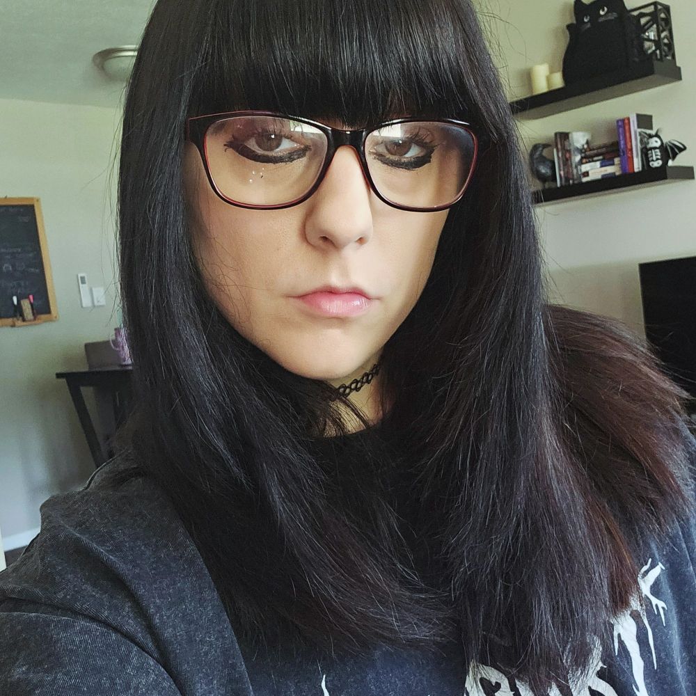 A selfie of Lex. She has long black hair and bangs and black glasses. She is wearing a big shirt that says "support your local library" in a death metal font.