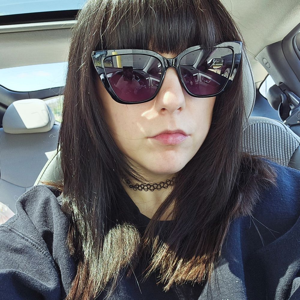 Lex in her car waring big cat eye sunglasses with purple lenses. Her hair is dark and is now layered to frame her face.