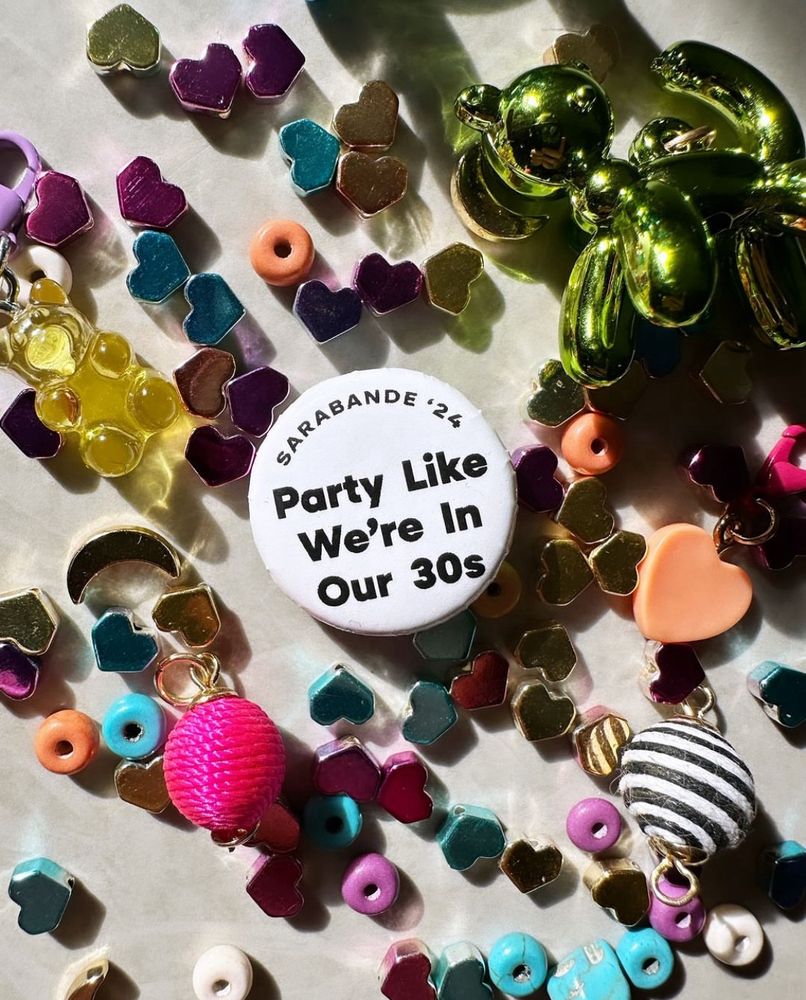 In a jumble of sparkly beads & baubles, a b&w pinback button: “Sarabande ‘24, Party Like We’re In Our 30s”