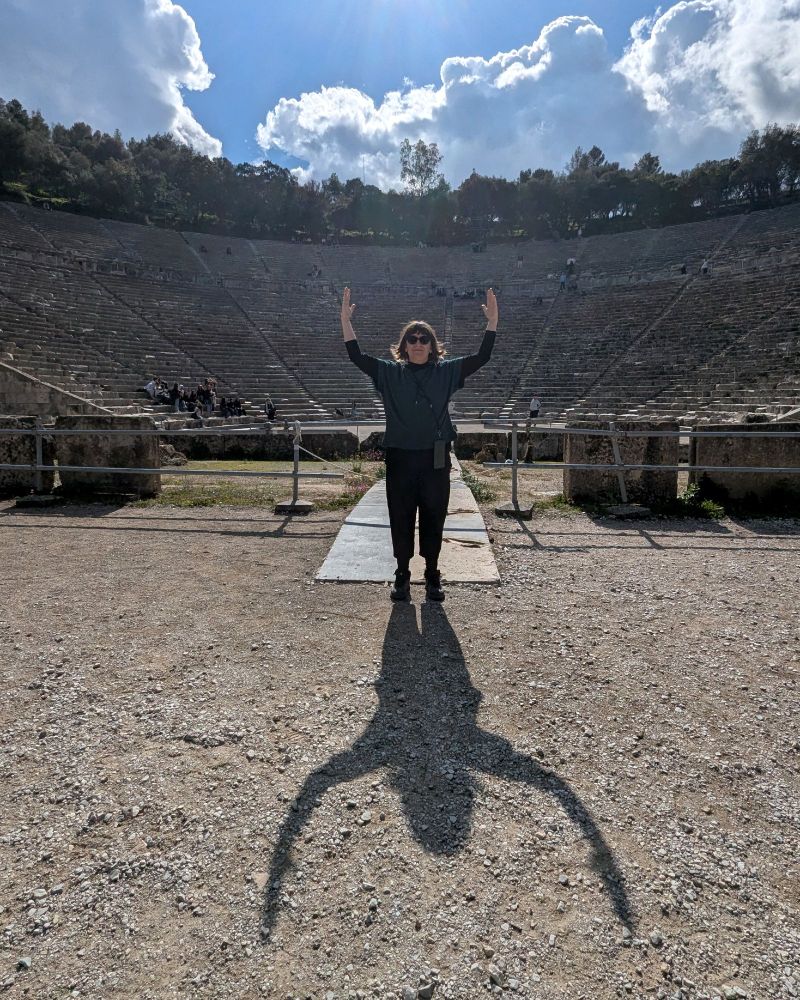 A woman with shoulder-length dark hair, wearing black sunglasses, a dark green sweater, and black pants, stands in the center of the ancient Epidaurus Theater at the Sanctuary of Asklepios in Greece. She has both arms raised towards the sky, with an expression of triumph or theatrical performance. The sun shines brightly behind her, casting a dramatic shadow on the gravel ground, which resembles the silhouette of a bird with outstretched wings. The massive stone seating of the theater rises behind her, partially filled with visitors scattered throughout. The sky is a mix of bright blue and fluffy white clouds, adding depth and contrast to the scene. A low stone railing separates the person from the seating area, and a narrow path leads directly to where they are standing.