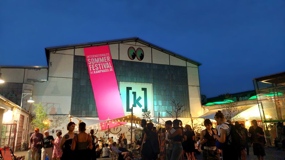 Evening scene at the Kampnagel International Summer Festival in Hamburg. A crowd of people is gathered outside under string lights and tents in front of the large industrial building with the [k] logo and a bright pink banner. The sky is dark blue, signaling dusk.