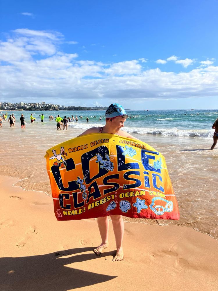 Woman standing barefoot on the sand at a beach next to the water holding a towel in front of her with Cole Classic written on it. She’s wearing a swimming cap and goggles on top. The sun is shining.