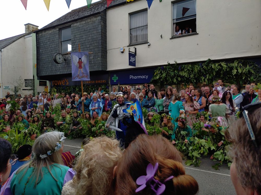 A picture taken on a street in Helston, costumed performers are enacting a verse from the Hal-an-tow. A man in Romanesque armour and welding a sword and shield (St Michael) faces down a purple garbed demon.