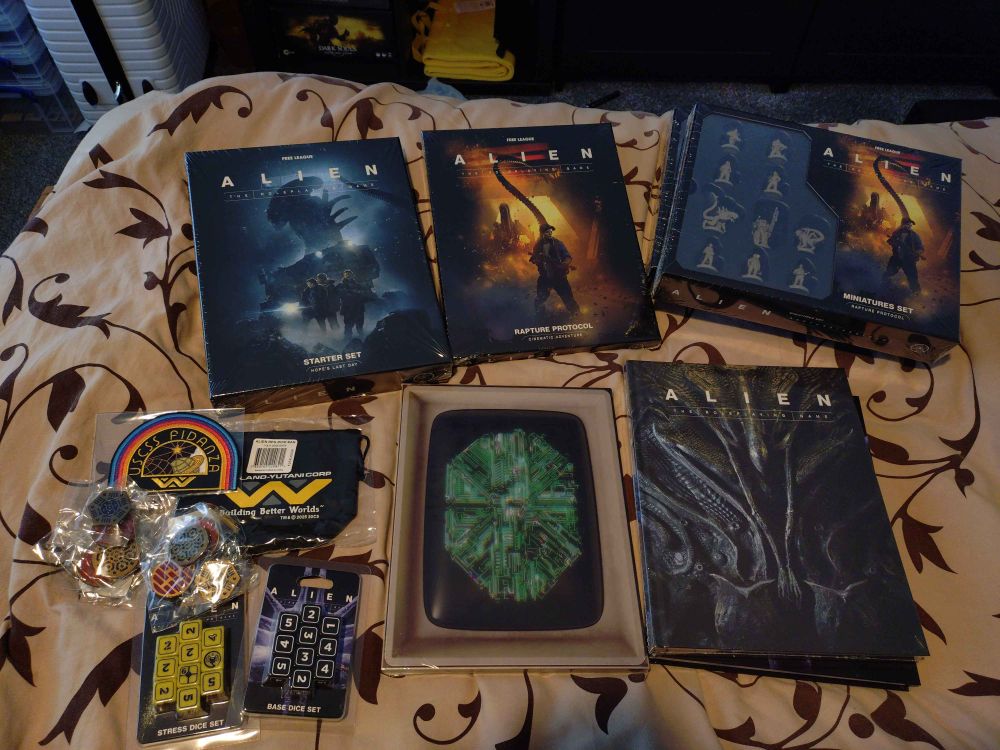 Assorted Alien RPG Evolved Edition products laid out on a bed. "Hope's Last Day" starter set, "Rapture Protocol" adventure, miniatures set, MUTHR's screen, Alien RPG Evolved Edition core rules, two sets of dice and some extra swag: a patch, dice bag and assorted pins.