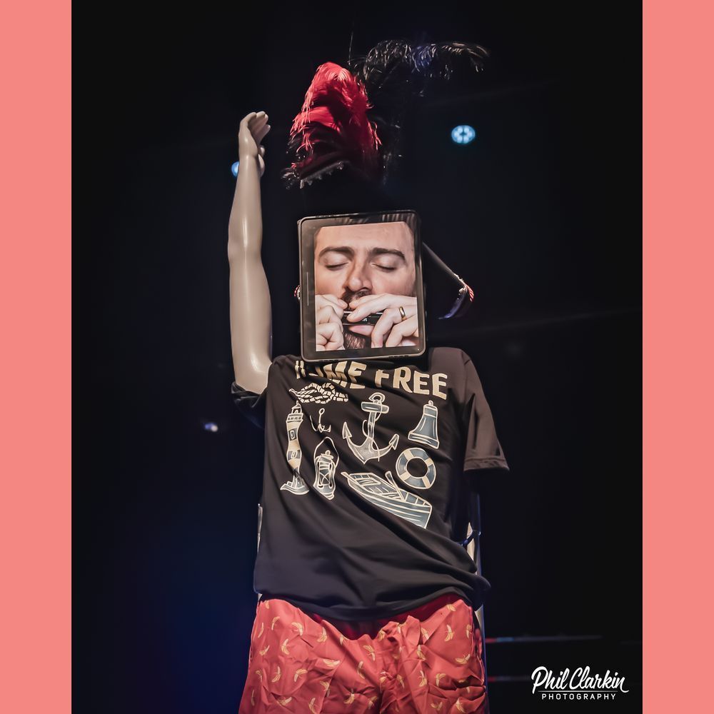 A picture of iRobbie. This is a mannequin wearing an ostentatious feathery hat, a Home Free tshirt, and orange shorts with.. croissants on them? The right arm is pointing straight up, perhaps in celebration. Then where the head should be, Robbie's face is visible with his own hands holding a harmonica up to his mouth.