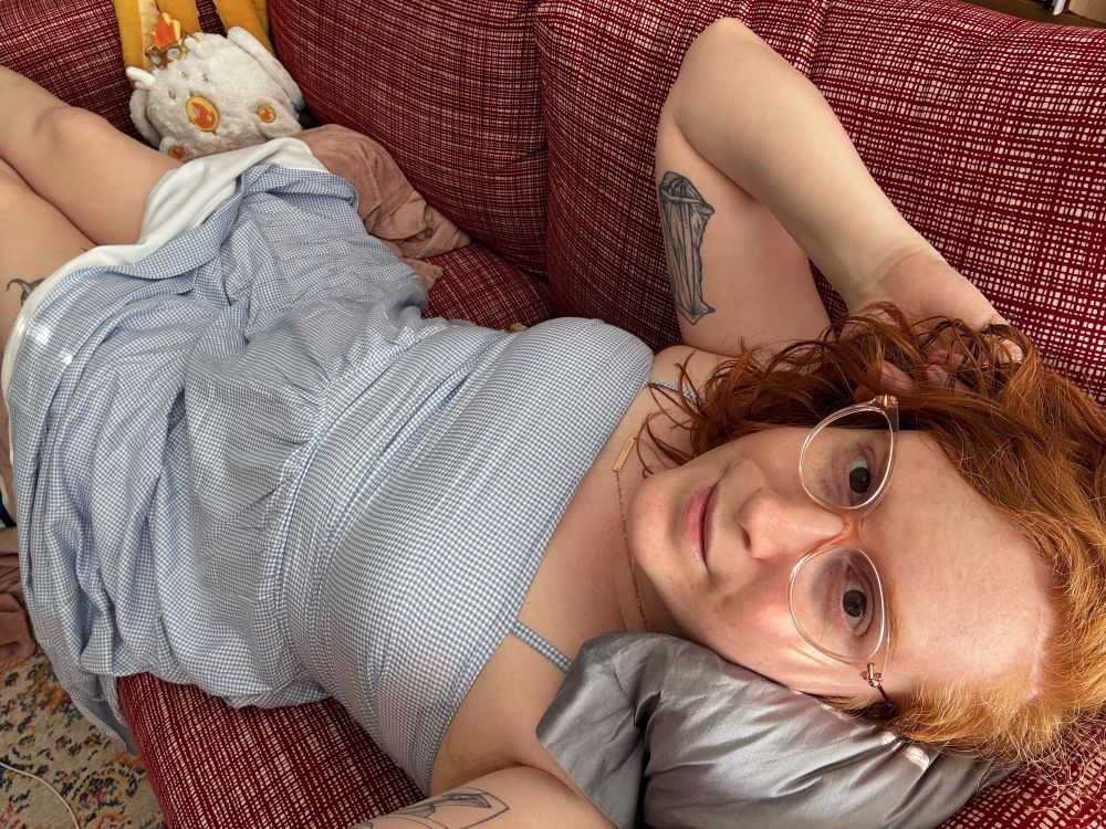 A long-haired femme redhead (Florence) on a red couch wearing a blue and white spaghetti strap summer dress