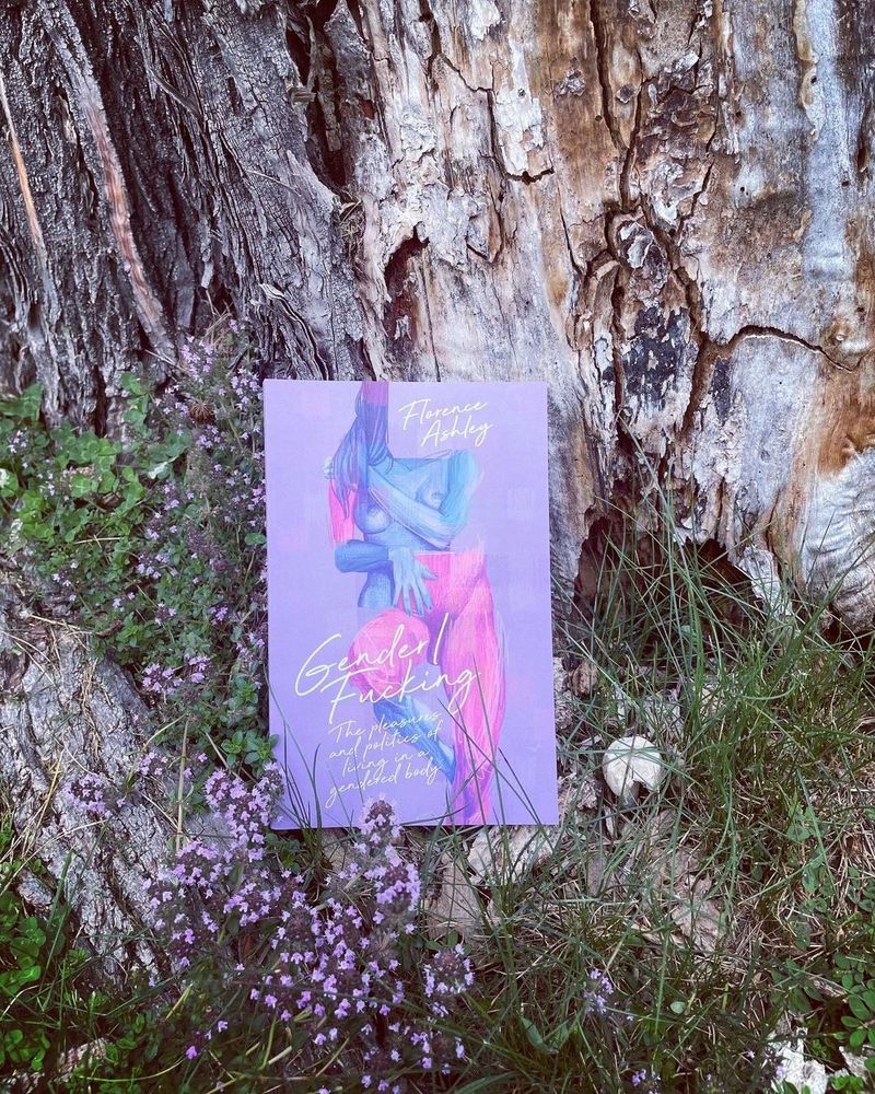 Copy of the book Gender/Fucking: The Pleasures and Politics of Living in a Gendered Body. The book is laying against a tree trunk with little flowers around it. The book cover is lavender with the top half of a naked character wih breasts and the bottom half of a naked character with a penis.