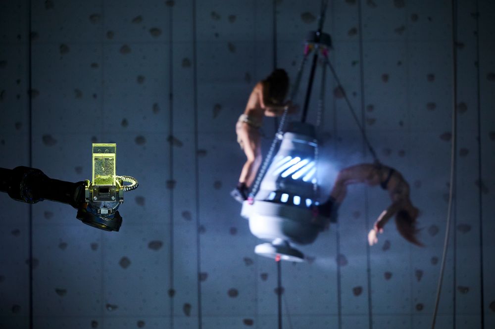 Bühnenfoto aus SANCTA: AUf einem riesigen, von innen beleuchteten und rachenden Weihrauchfass sieht man im Hintergrund zwei nackte Performerinenn stehen, eine von ihnen nach hinten gebeugt. Im Vordergrund ein Roboterarm, der einen kubischen Behälter mit einer durchsichtigen Flüssigkeit hält. Foto: Matthias Baus