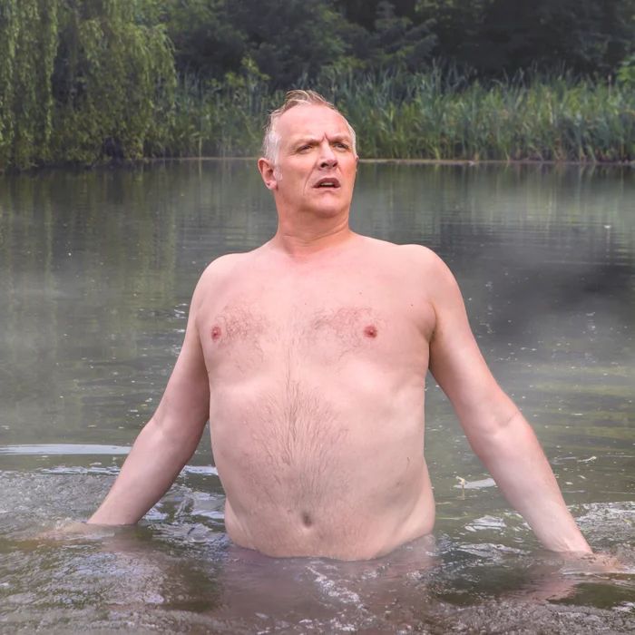 Greg Davies standing waist deep in a pond bare chested looking at something in the distance. 