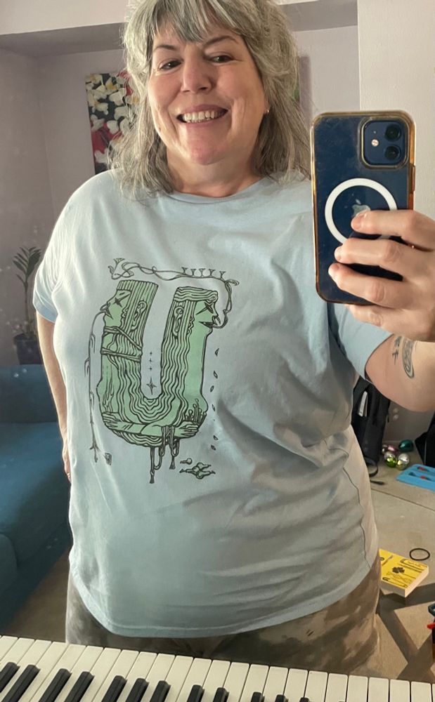 A woman with gray hair wearing a light blue t-shirt with an interesting design of witch heads connected by a u-shaped tube. 