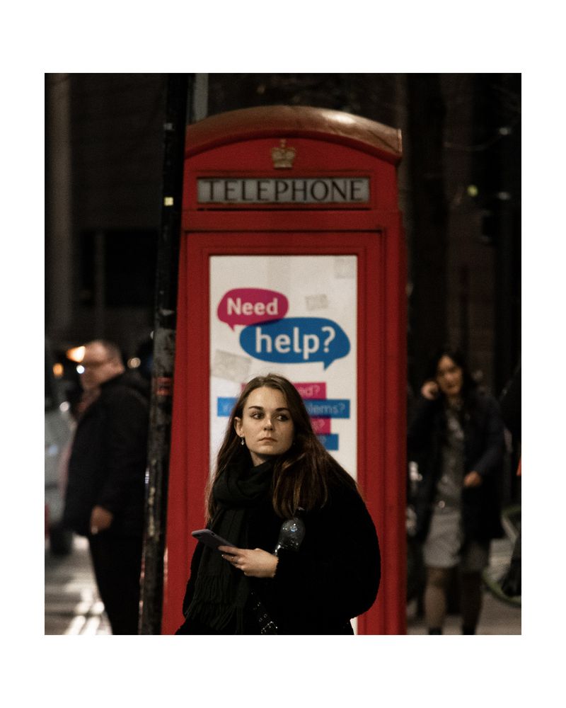 Candid street photo from London of a woman standing in front of a red phone booth. 