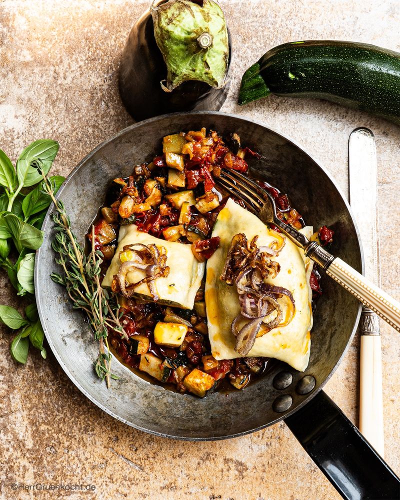 Hellbrauner Steinhintergrund. Darauf eine kleine, runde, silberne Pfanne. Darin italienische Maultaschen mit Auberginen, Zucchini und weiterem Gemüse gefüllt.