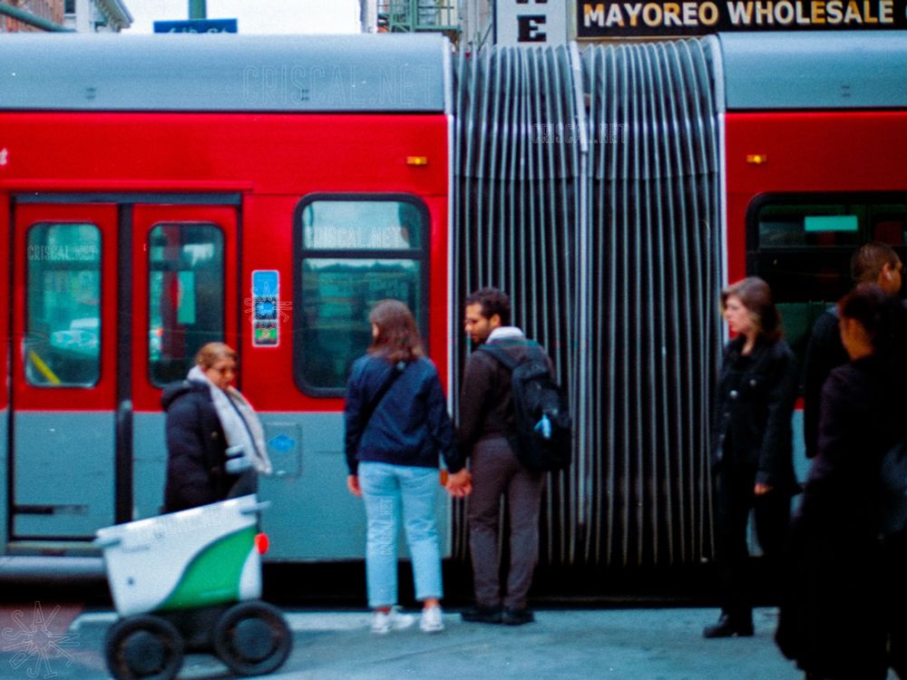 A red city bus is stopped on a busy street as pedestrians wait nearby. A couple holding hands stands in the foreground, while others pass by. A small white and green delivery robot rolls past on the left side of the frame. The scene captures the movement and mix of people observing the robot. #photography