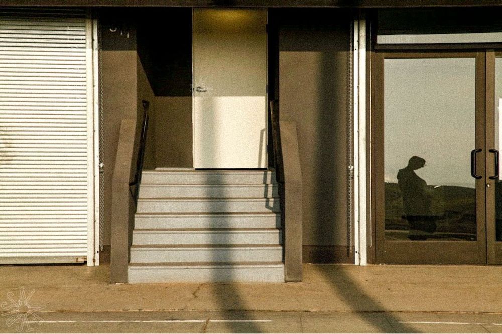 A minimalistic exterior scene featuring a beige building with a small staircase leading to a door. To the right, a reflective glass door shows the shadowy silhouette of a person holding something, possibly a phone. #photography