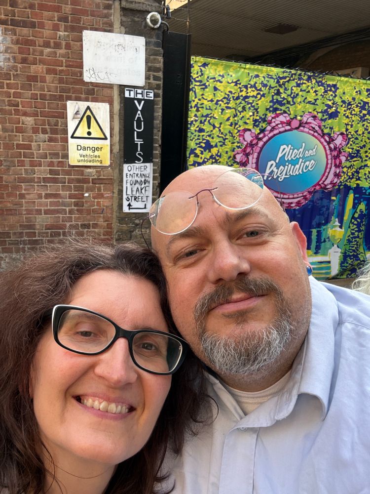 Man and woman in front of a sign “plied and prejudice”
