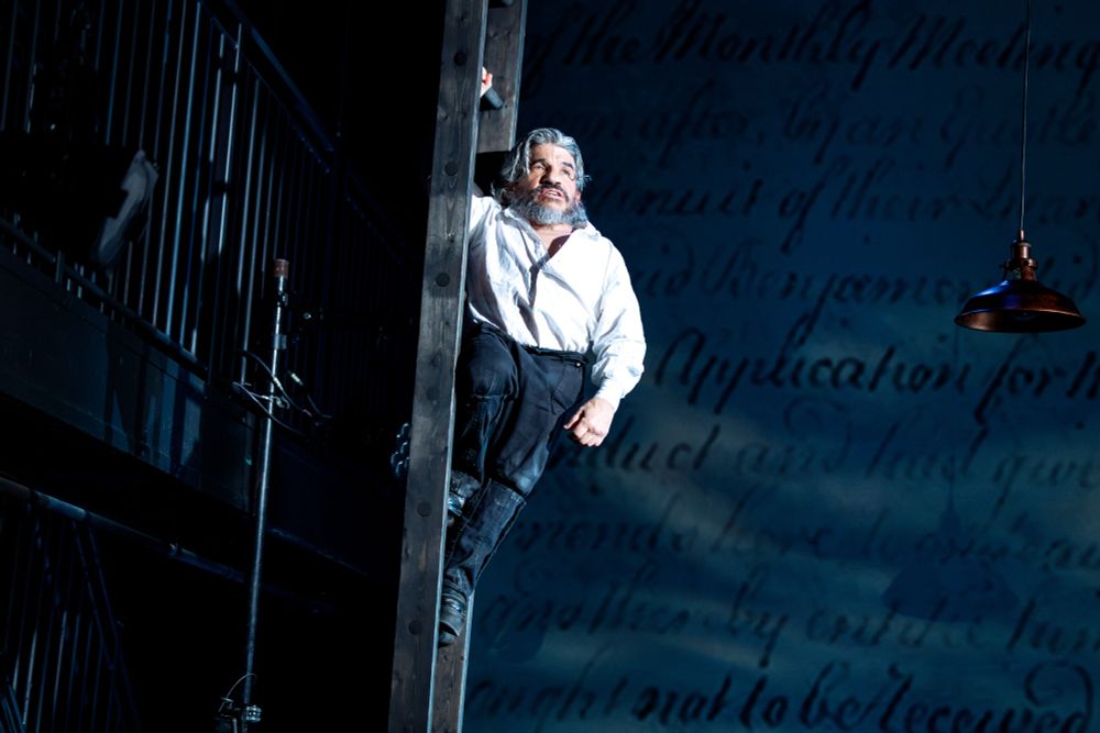 Dressed as Benjamin Lay, Mark Povinelli holds onto a ladder. The stage is washed in blue, evoking the feeling of being on the water.