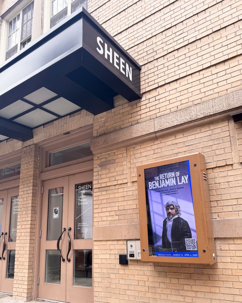 We see the outdoor black awning with the word, “SHEEN,” in all caps. Underneath the awning, we see a vertical board with the poster for THE RETURN OF BENJAMIN LAY, featuring an image of Mark Povinelli in character. 