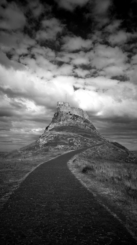 A moody black and white shot of  Lindisfarne Castle, with a pathway in the foreground, and a rolling field of clouds above.
