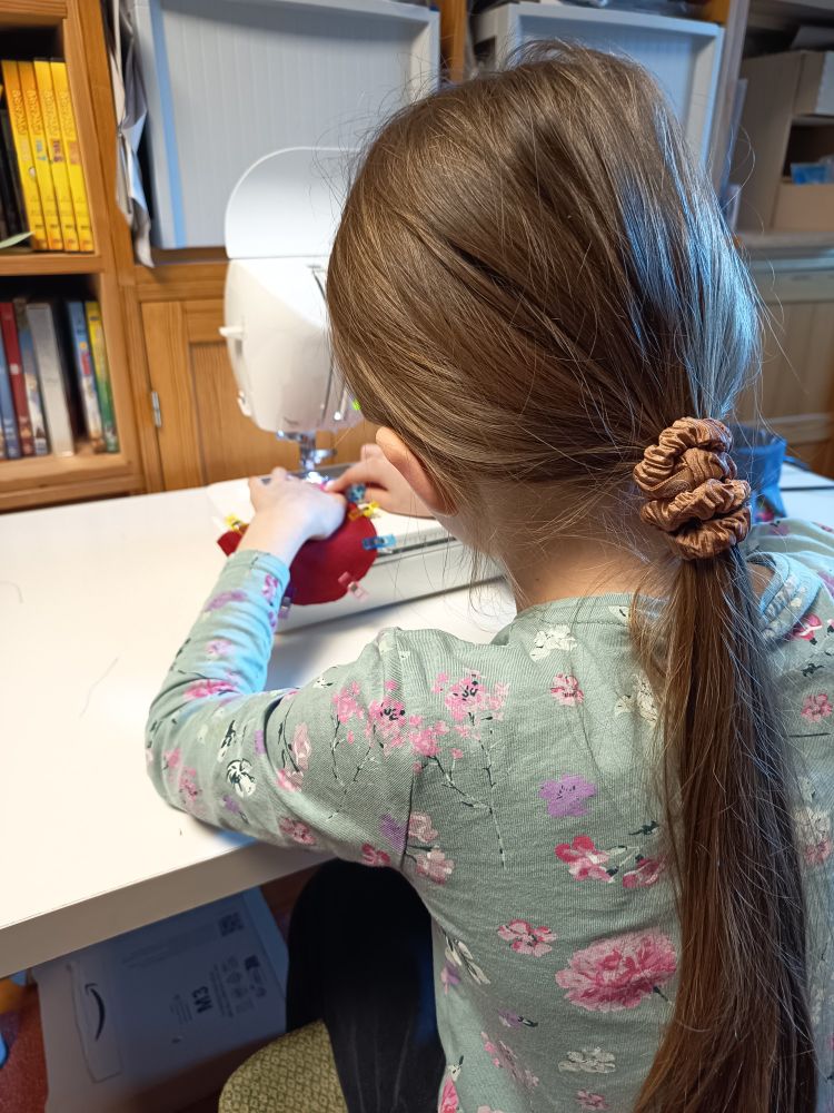 Kind von hinten zu sehen, mit blonden langen Haaren, zum Zopf gebunden, sitzt vor einer Nähmaschine und näht ein rotes Stück Stoff zusammen