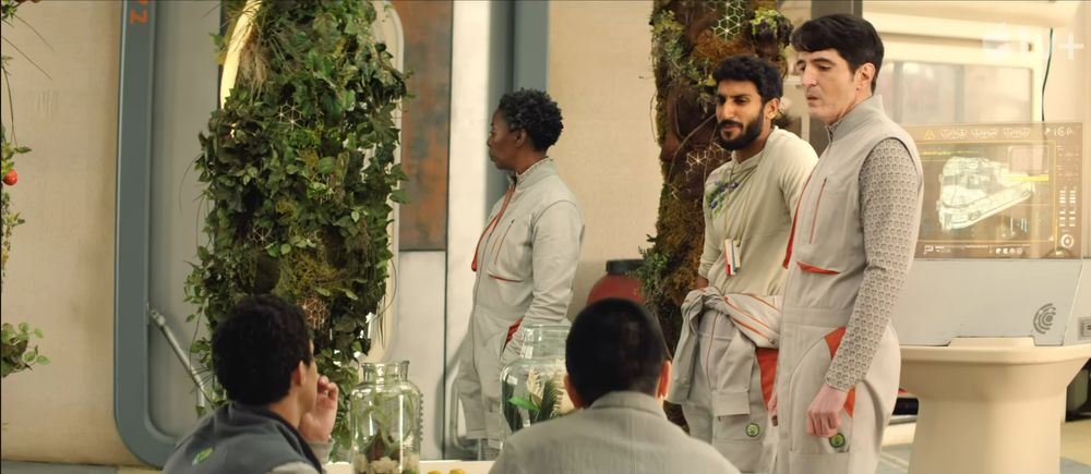 The space hippies inside the habitat.  Their clothing is a mix of soft greys, red, tan and one dark grey.  There are plants around the supporting coloumns, and the future is a light tan as well.  In this shot Mensah is standing and looking away from the camera.  Gurathin and Ratthi are also standing and looking towards Pin-Lee and Arada who are sitting and looking at them