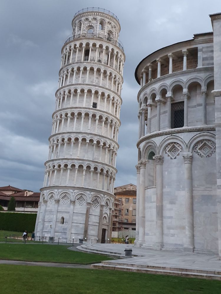 The leaning tower of Pisa.  A white stone building leaning at a slightly alarming angle.  It looks a lot like a wedding cake with 8 clear levels, 6 of which have columns on the outside.  There is another, shorter, white stone building in the foreground of the image, made in a very similar style with columns as decorations.  The grass of the area is very green, there are a few people visible, and behind it all is a grey cloudy sky.