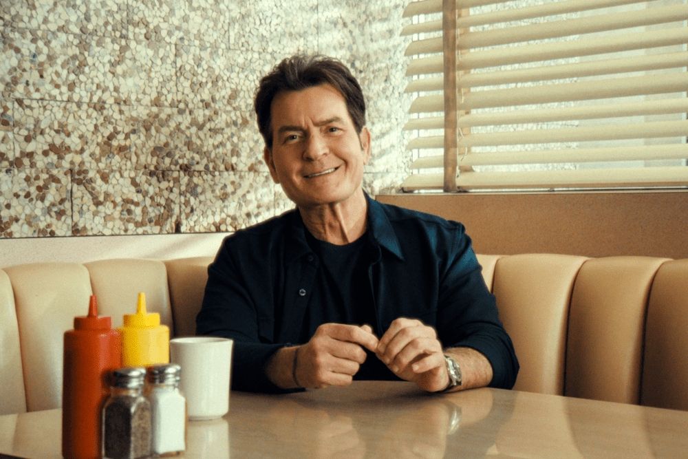A beaming Charlie Sheen sitting at a diner booth, staring at a camera pointing at him. With a red and yellow ketchup and mustard bottles, along with salt and pepper bottles. He's drinking coffee. From the documentary AKA CHARLIE SHEEN. 