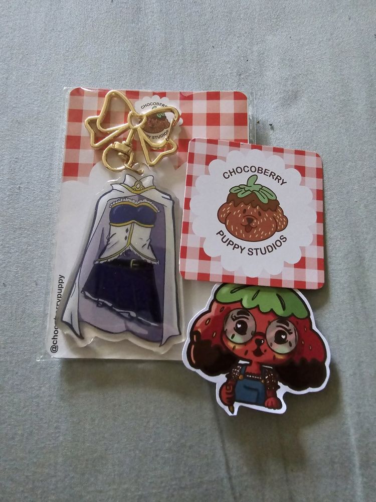 On the left you see a bow shaped keychain with the outfit of the character Sayaka from Madoka Magica. Her blue and white corset and blue skirt with white cape. 
On the right you see a puppy depicted as a strawberry on a card. The text on it says: Chocoberry Puppy Studios
Underneath that is a sticker of that same puppy in a more anime style. 