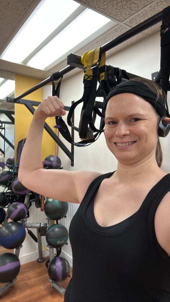 White woman in black tank top and headband forces her right arm while smiling at the camera. Rack of medicine balls behind. 