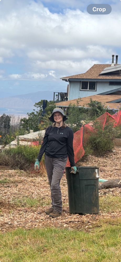 White woman in dark clothes and blue gloves stands next to a trash bin full of mulch. A blue house is behind her on the right. Ocean and mountains view on the left. 
