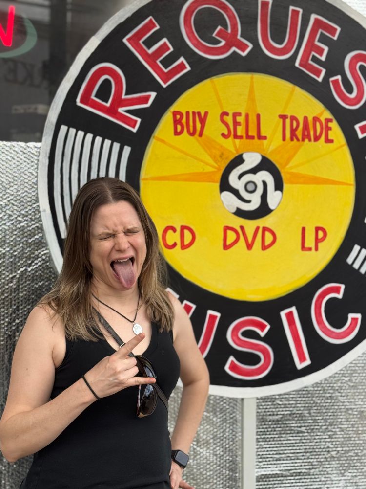 Blonde, white woman sticks out her tongue and does the horn gesture with one hand. Eyes shut and tongue sticking out. Standing in front of a hand painted sign with “Request Music” “CD DVD LP”