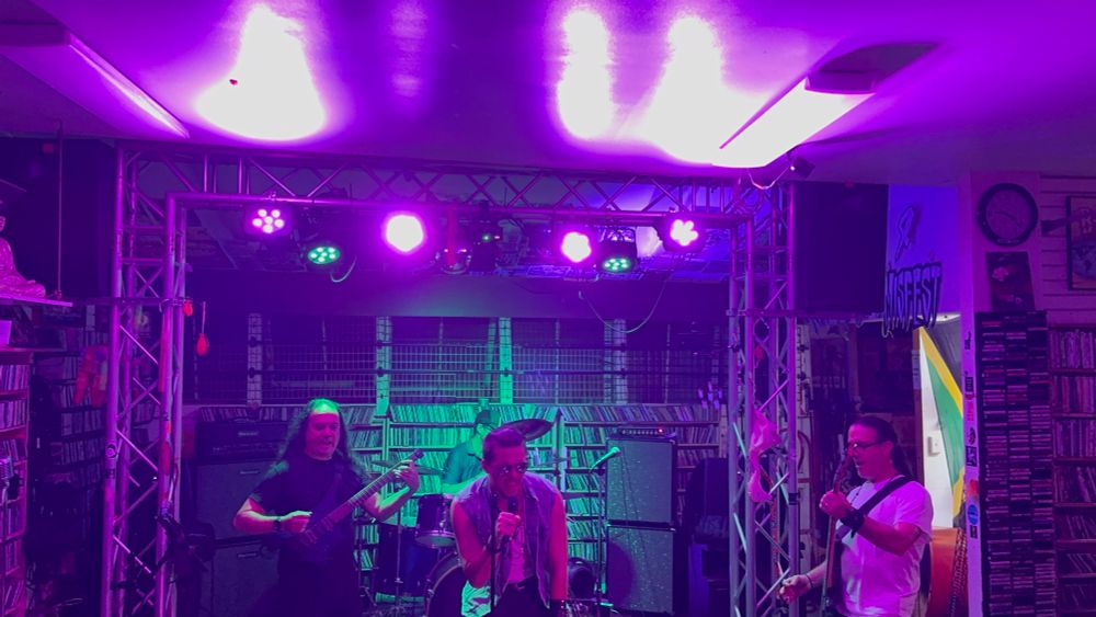 Four piece rock band performing with stage lights above and CDs along the walls. 