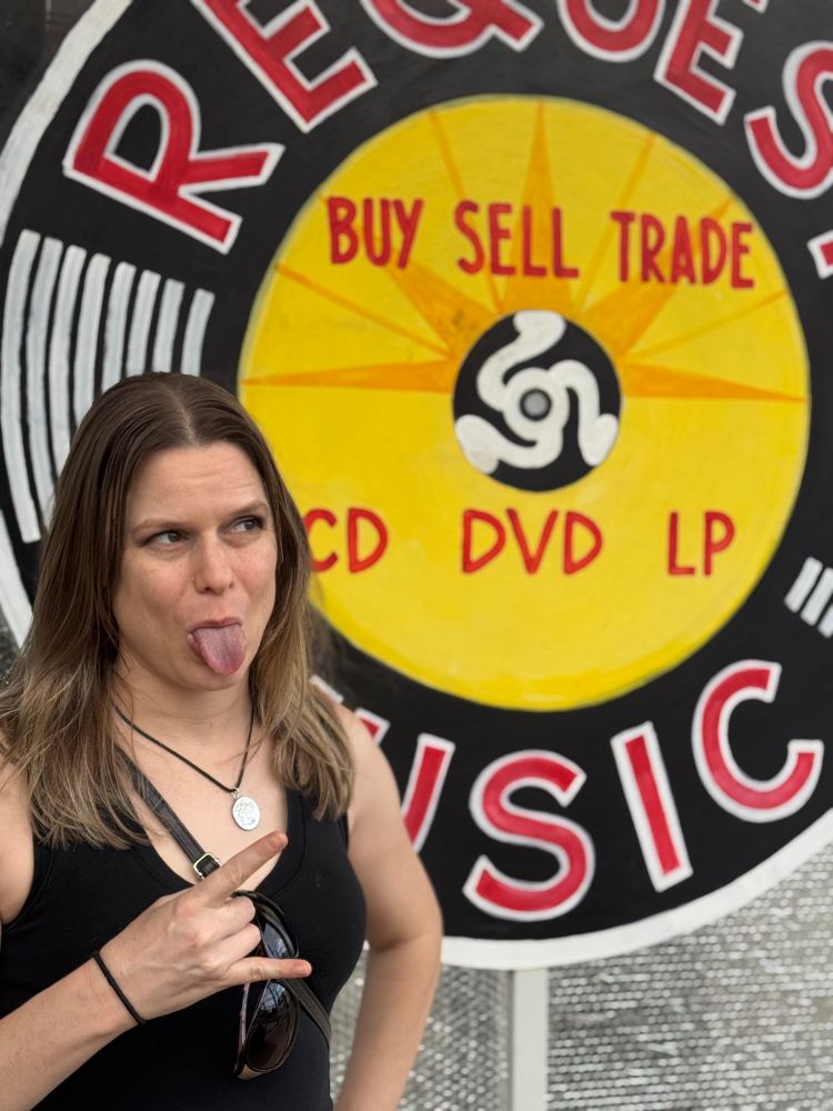 Blonde, white woman sticks out her tongue and does the horn gesture with one hand. Standing in front of a hand painted sign with “Request Music” “CD DVD LP”