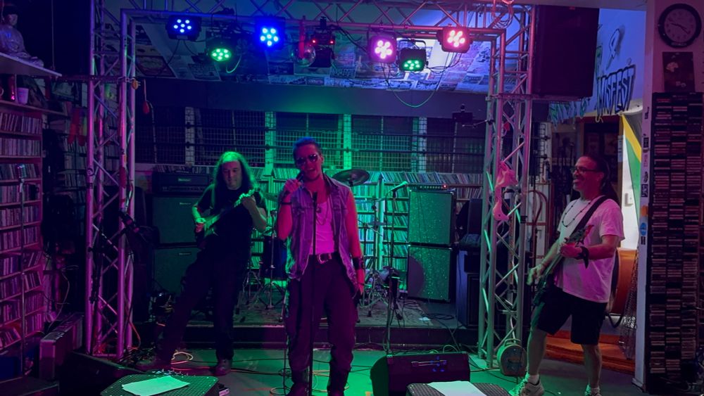 Four piece rock band performing with stage lights above and CDs along the walls. 