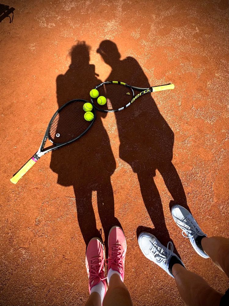 Schatten von 2 Personen auf einem Tennisplatz, auf dem Boden liegen 2 Tennisschläger und Tennisbälle