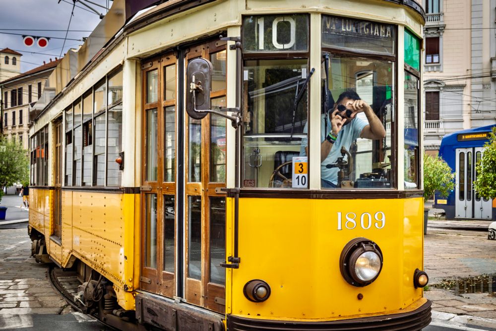 Der Führer einer historischen Straßenbahn sitzt in der Fahrerkabine und hält eine fiktive Kamera in der Hand als würde er die Person, die ihn fotografiert, ebenfalls fotografieren. 