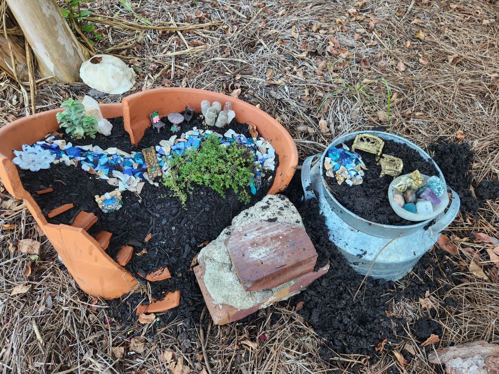 Fairy garden built from a broken clay pot and a metal planter. In the foreground are bricks from an old foundation. Many tiny, fairy-sized furniture, plants, and decorative elements on the top.