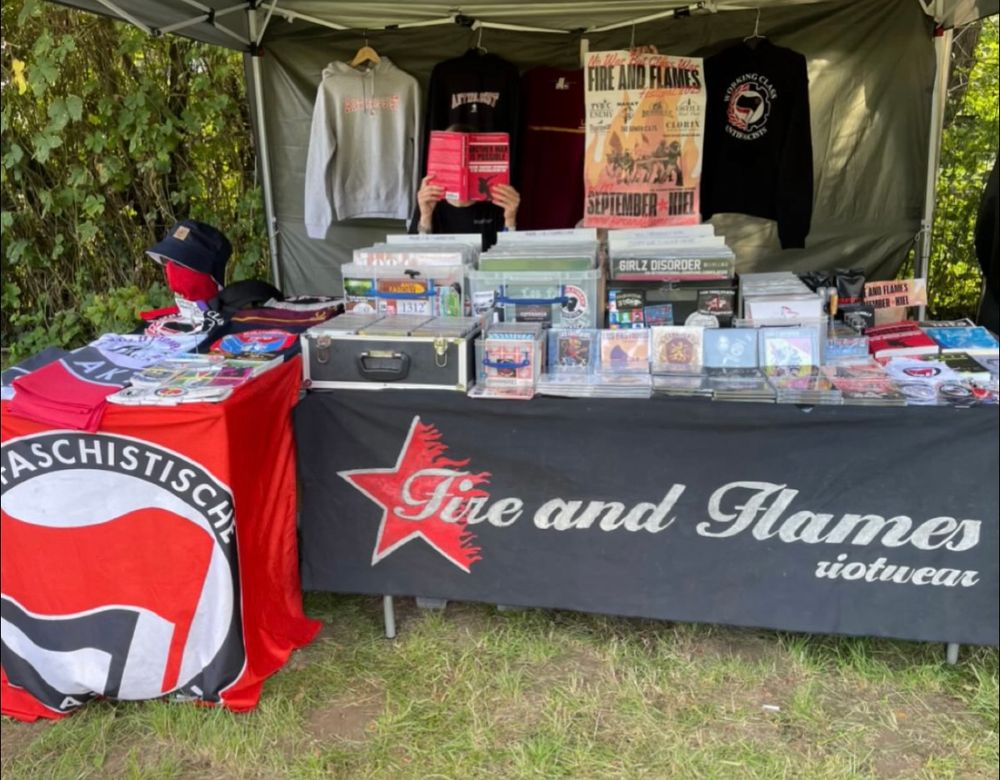 Fire and Flames Music merch table, where records, books, and shirts can be seen. A person behind the table conceals their face with a copy of ANOTHER WAR IS POSSIBLE.

Front of table is covered with a large FIRE AND FLAMES RIOTWEAR banner and an ANTIFASCHISTISCHE AKTION flag.