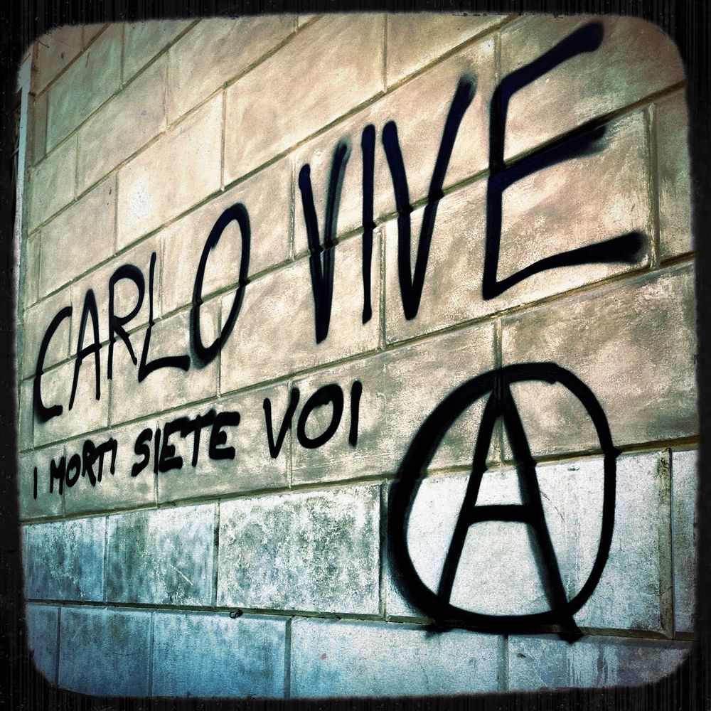 Graffitti on a brick wall in black letters reading CARLO VIVE, I MORTI SIETE VOI and a circle A.