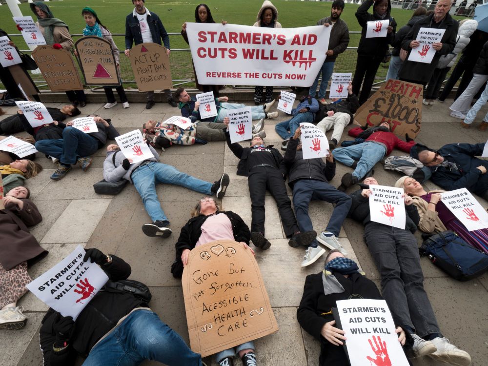A group of activists stage a 'die-in' protest outside of Parliament. They hold posters and gravestone-shaped placards that say 'Starmer's aid cuts will kill'