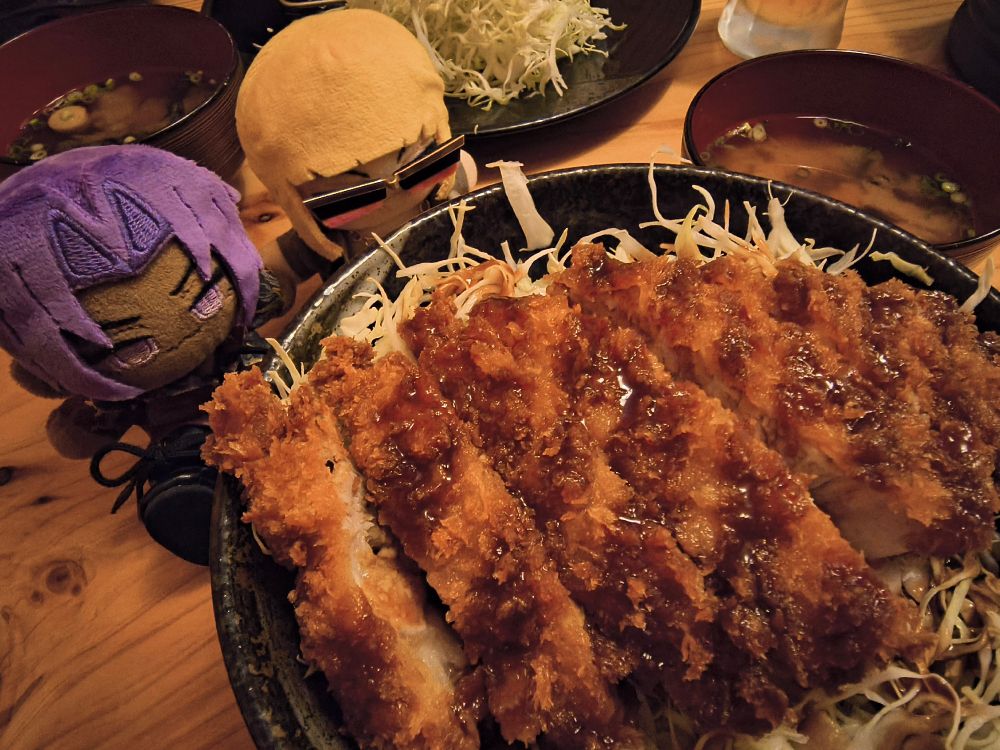 画面手前に千切りキャベツのうえにとんかつがのったどんぶり、その左奥側にぬいぐるみが2体置かれており、右側奥にみそ汁の碗がある。