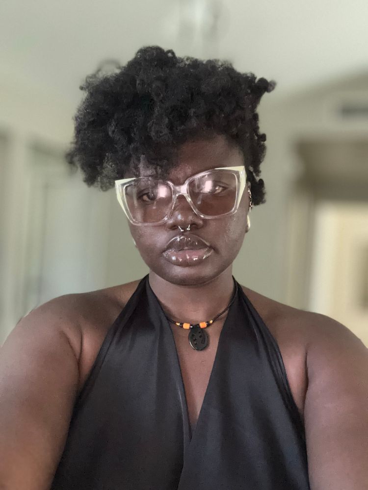 A black person with a Mohawk Afro wearing a septum, a gye nyame pendant and shades.
