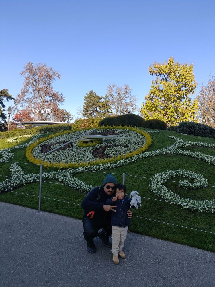 Galean y su sobrino frente al reloj de flores de Ginebra. Galean tiene lentes oscuros y está en cuclillas al nivel de su sobrino haciendo el signo de paz. El sobrino saca la lengua.